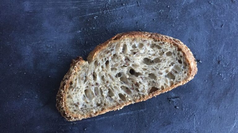 A beautifully golden-brown, crusty no-knead artisan bread loaf cooling on a wire rack, with a Dutch oven in the background, s...