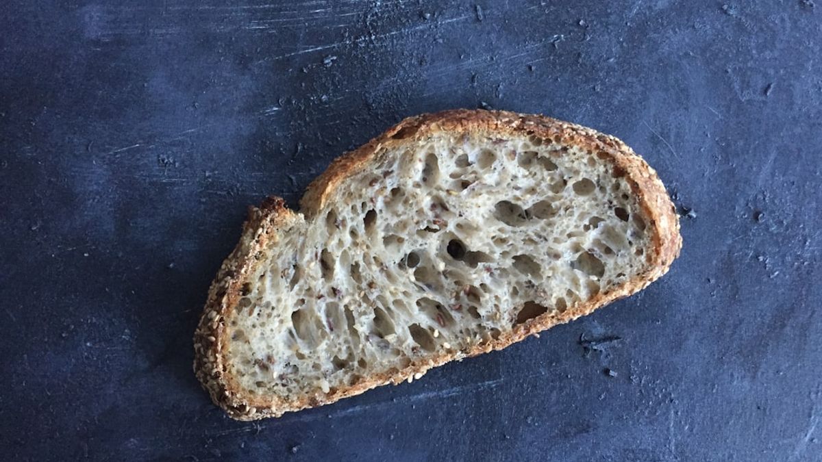 A beautifully golden-brown, crusty no-knead artisan bread loaf cooling on a wire rack, with a Dutch oven in the background, s...
