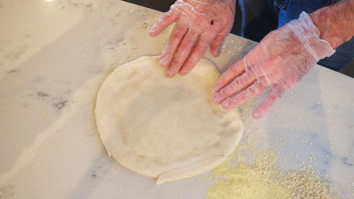 Close-up of hands gently stretching homemade pizza dough on a floured wooden surface, with a dusting of flour and a rustic ki...