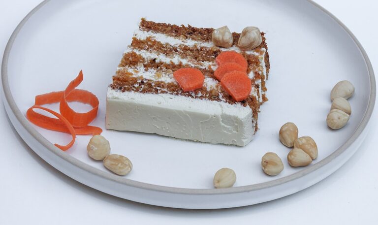 Two-layer carrot cake with cream cheese frosting on a white cake stand with walnut garnish and a slice removed to show moist interior