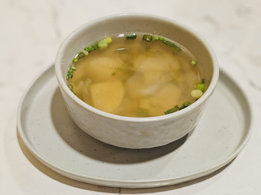 A pale golden broth in a white bowl with a bay leaf floating on the surface