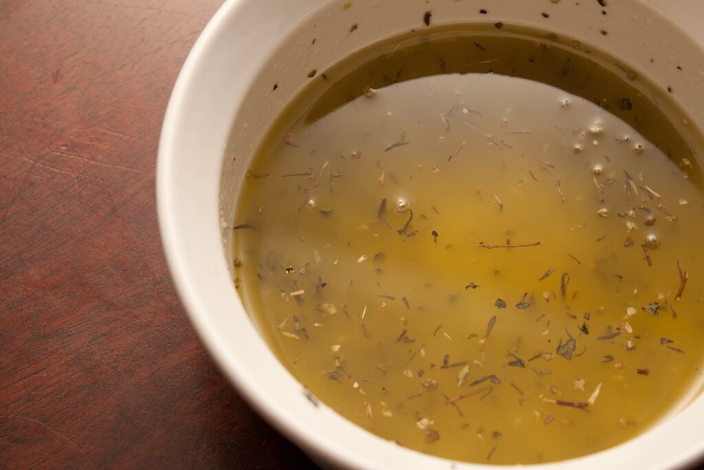 A small jar of golden vinaigrette beside a whisk and a bowl of green leaves