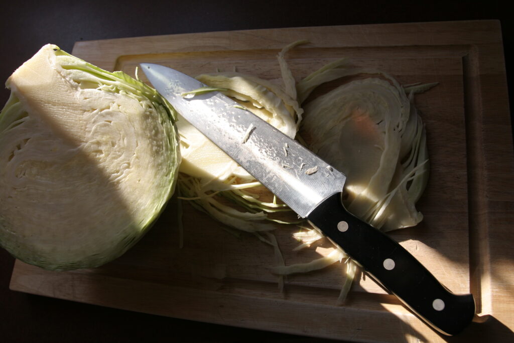 A halved white cabbage on a wooden board beside a long knife and fine shreds