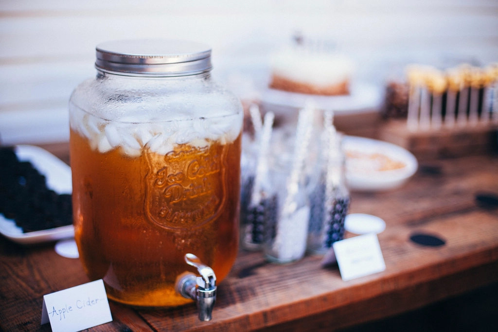 Glass bottles of cloudy apple cider standing on a wooden shelf beside fresh apples